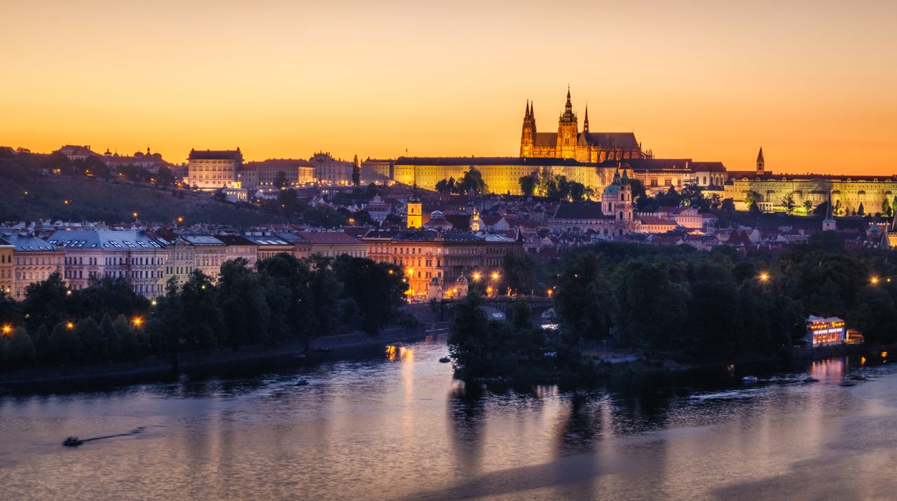 Scenic view of Prague Castle glowing at sunset with the Vltava River in the foreground.